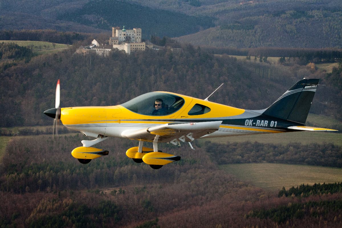 Yellow Bristell Classic in flight past a hilltop castle